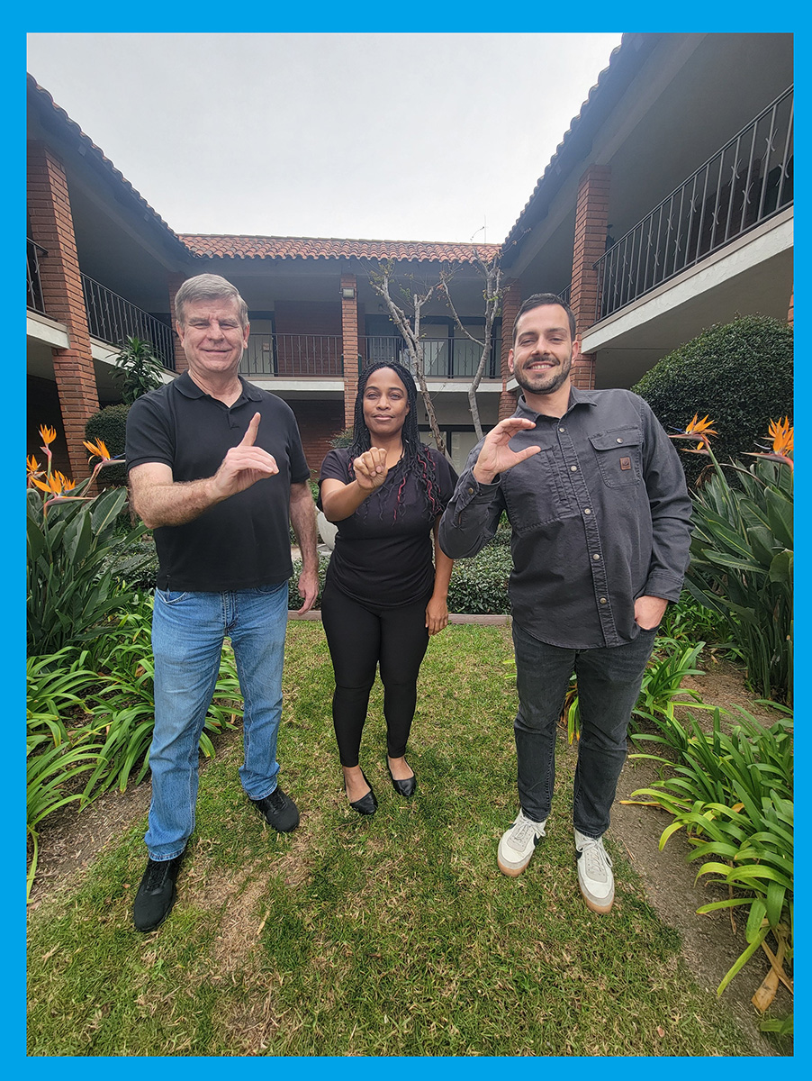 Pictured above: Deaf Services Team Jet, Pamela and Tobias signing D-M-C in ASL standing outside in courtyard with Bird of Paradise orange foliage behind them