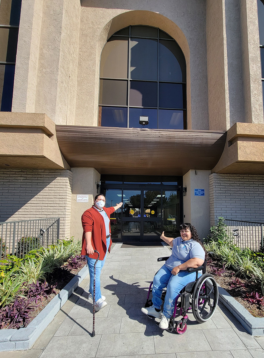 DMC Youth Connect Team, Luz and Nelly saying “Welcome to College” pointing toward college doors as they smile outside building on ramp