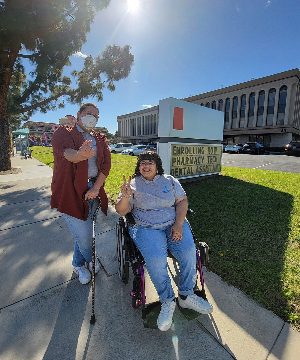 DMC Youth Team Luz and Nelly outside by now enrolling sign on sidewalk with sun shining and trees in background near grass and bulding 
