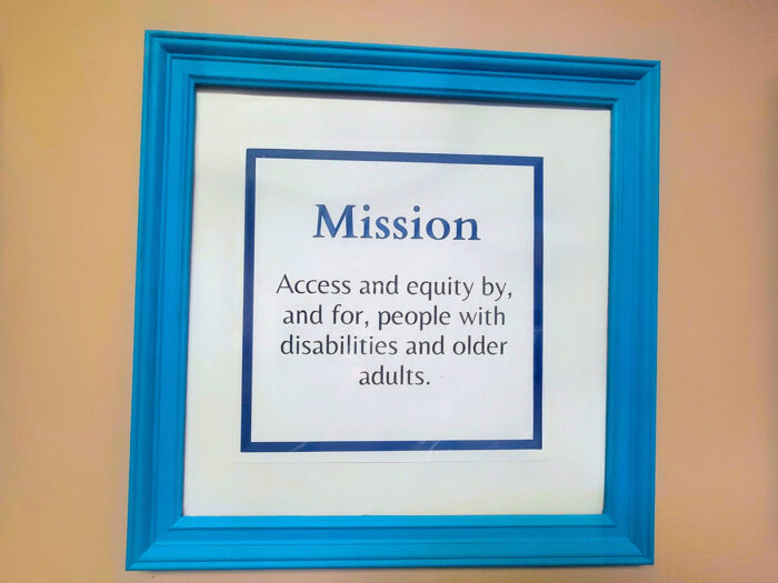 Photo of DMC’s Mission Statement in blue frame on tan wall “Mission Access and equity by, and for, people with disabilities and older adults