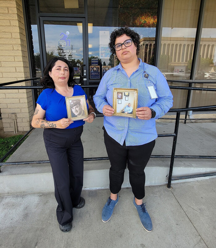 L-R: Marysol Cuadrado, Lead VIP Advisor holding photo of late Veteran DMC second DMC Board President, Mick Spencer in frame and Michelle Ramos, Director of Community Support Services holding photo of original DMC Board President, Greg Winterbottom in gold frame standing outside DMC building looking somber