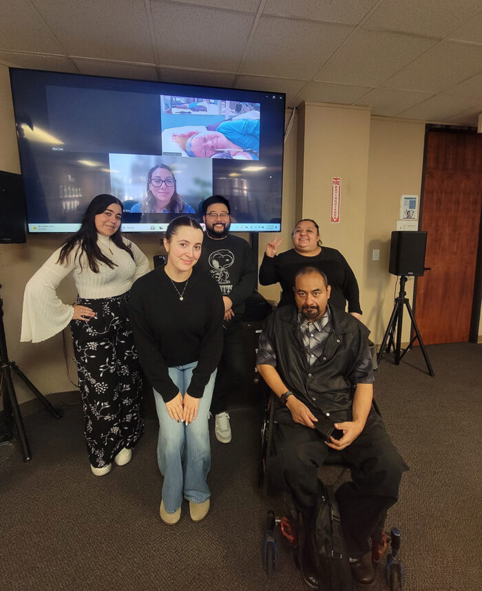 BreeAnna Adner, DMC Systems Change Advocate with other Southern California Systems Change Advocate in person (and digital on screen behind group) in regional meeting at DMC offices smiling.