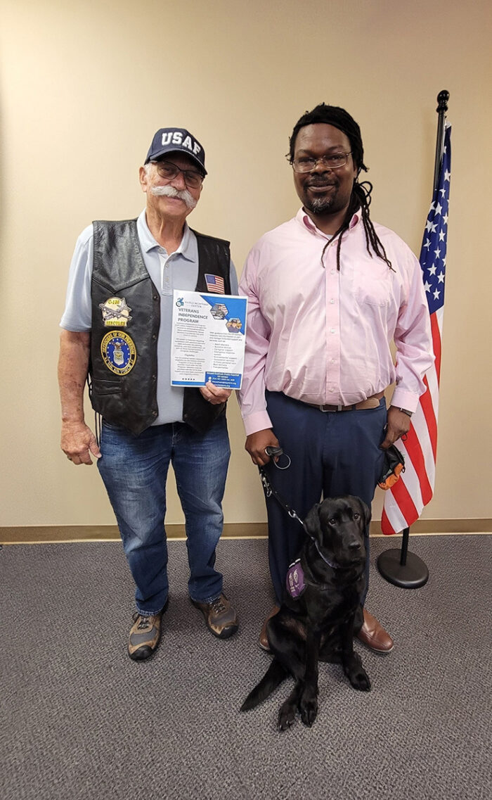 Veteran Tony Clement, DMC Board President and Veteran Griffin Dooley, DMC Board Governance with service dog, Vivy standing next to U.S. flag inside DMC offices smiling as Tony holds Veterans Independence Program flyer