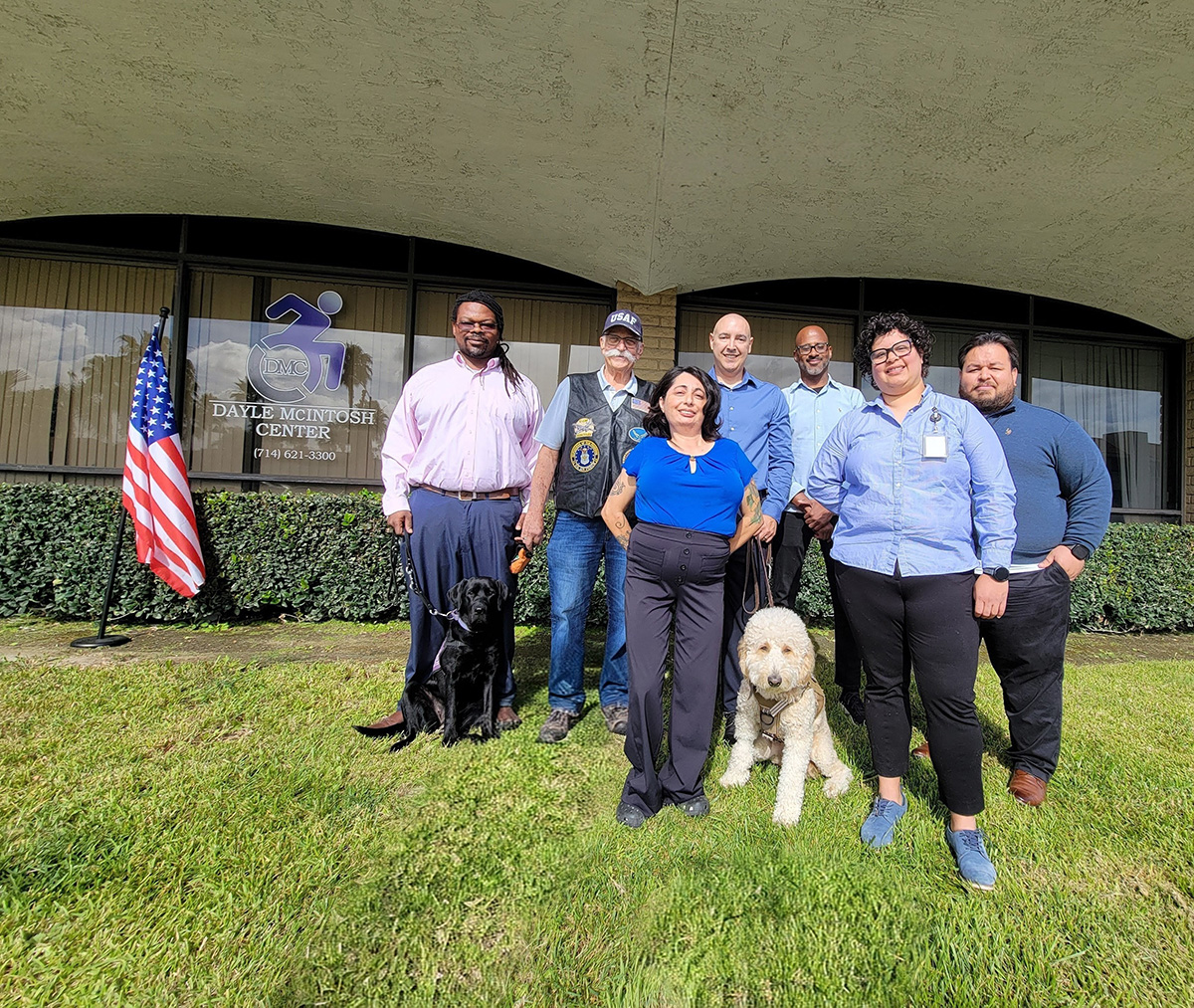 Group photo in front of DMC offices on lawn with U.S. flag in the background. Front row L-R: Marysol Cuadrado, Lead VIP Advisor, Michelle Ramos, Director of Community Support Services. Back row L-R Griffin Dooley, DMC Board Governance Chair with Service Dog Vivy, Tony Clement, DMC Board President, Eric Rodriguez, VIP Advisor, Riverside with Service Dog Honey, Andrew Toney, VIP Advisor Los Angeles and Luis Alvarez, DMC VIP Advisor OC/L.A.