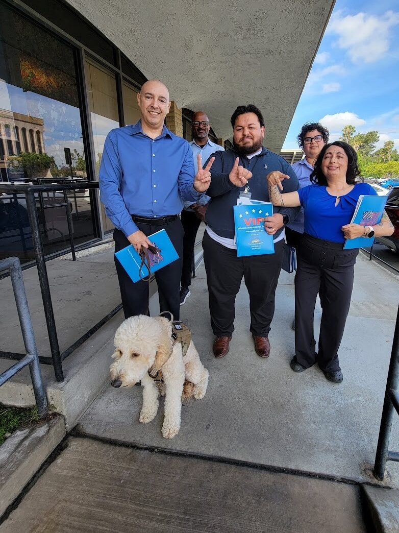 DMC VIP Team on ramp outside building with blue sky and white clouds on sunny day in background. L-R (Front Row) Eric Rodriguez, VIP Advisor, Riverside with “Honey” Service Dog; Luis Alvarez, VIP Advisor OC/LA, Marisol Cuadrado, Lead VIP Advisor – the three of them in the front row signing V-I-P in finger spelling ASL. Back row: Andrew Toney, VIP Advisor Los Angeles and Michelle Ramos, DMC Director of Community Support Services.