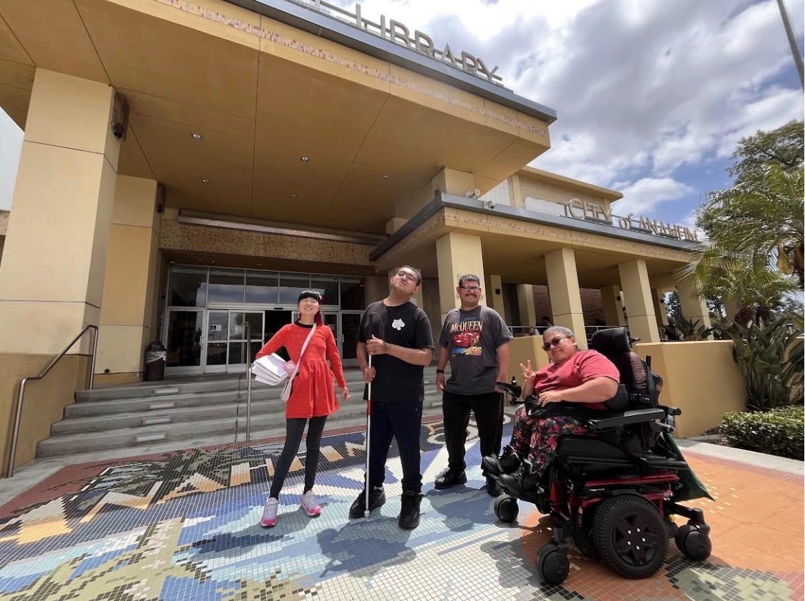 A group of cross-disability youth posing for a photo in front the library. They are smiling and doing poses. In the background, there are signs for “Library” and “City of Anaheim.”