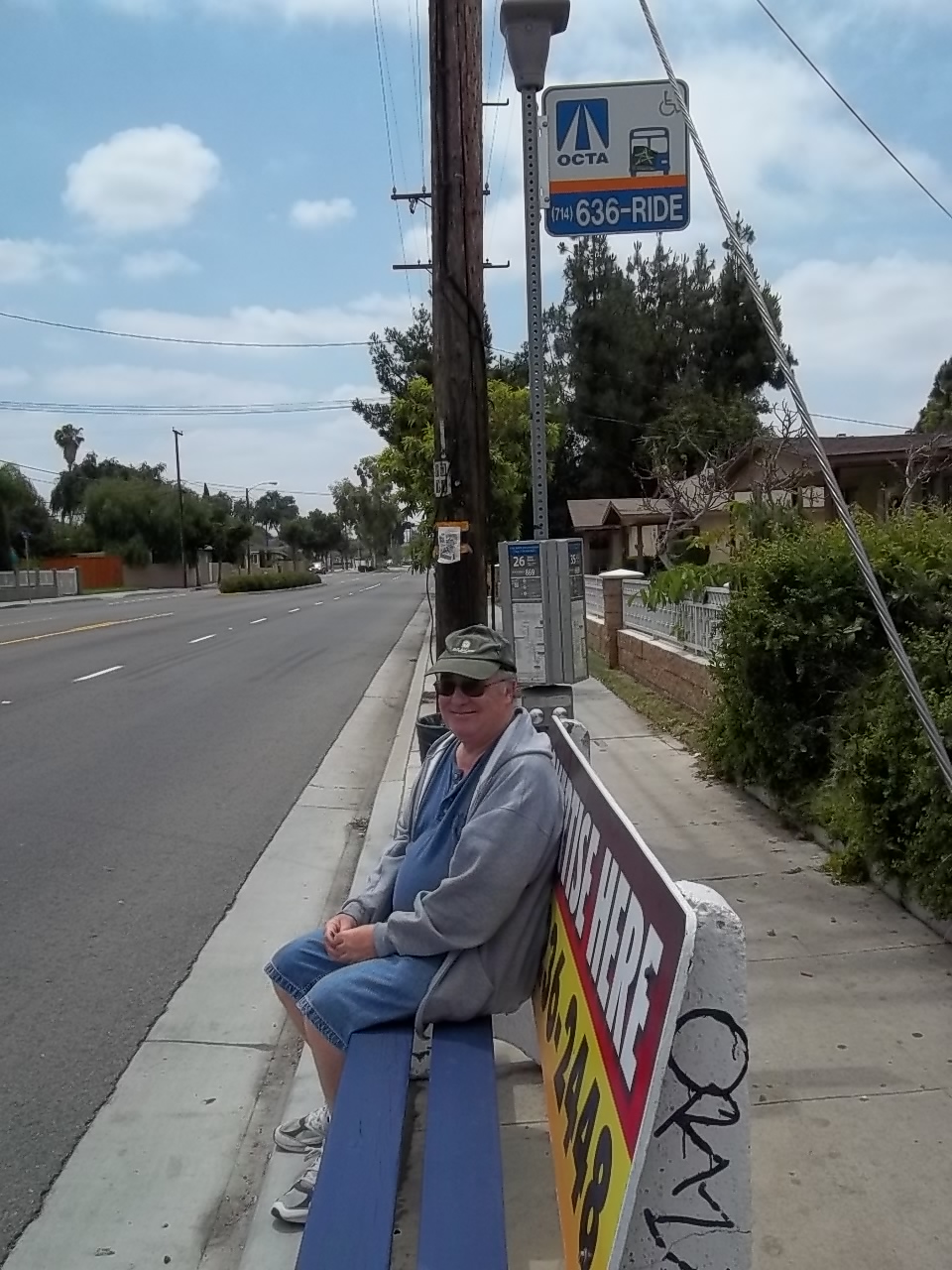 An older adult consumer smiling while he waits on a bench at a bus stop.