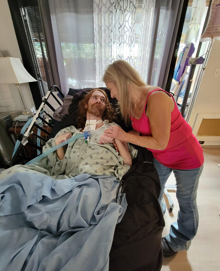 Transition consumer in his hospital bed with mother and primary caregiver at his bedside, holding his hand, and looking into his eyes lovingly.