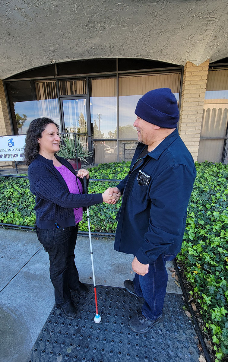 Gaby, DMC’s ILS Instructor for the Blind with consumer shaking hands outside as consumer begins journey with DMC for support. Both are smiling as consumer wears blue coat and matching ski cap, Gaby holds mobility cane wearing violet blouse as they stand at base of ramp outside DMC building.