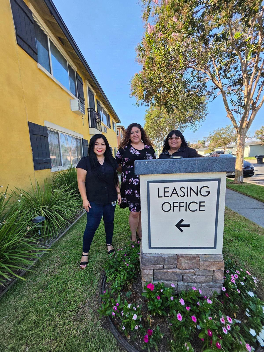 Housing team L-R Monica, Marisol and Paola with leasing sign featuring arrow to the left with pink flowers and shrubs at base, outside of yellow-painted apartment building 