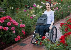 Picture-of-woman-in-wheelchair-with-tote-on-her-lap-rolling-on-pavers-through-a-rose-garden.-support services