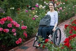 Services Picture of woman in wheelchair with tote on her lap rolling on pavers through a rose garden.