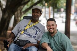 Peer Support Man in power chair with another bent down next to him.