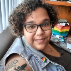 Woman with curly hair wearing glasses and a rainbow pin.