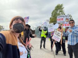 DMC’s Systems Change Advocate, Beck, smiling with a black KN95 mask on and making a peace sign with their left hand. They are a white person wearing an orange beanie, honey bear earrings, a brown and orange cardigan, and a shirt with corgis on it. Behind them, six ACCESS bus drivers stand with protest signs reading “Fighting for a FAIR contract” and “ON STRIKE AGAINST Transdev; Worker’s rights are human rights.” Several of them are making the peace sign as well and one of them is making the hand symbol for “hang loose.”