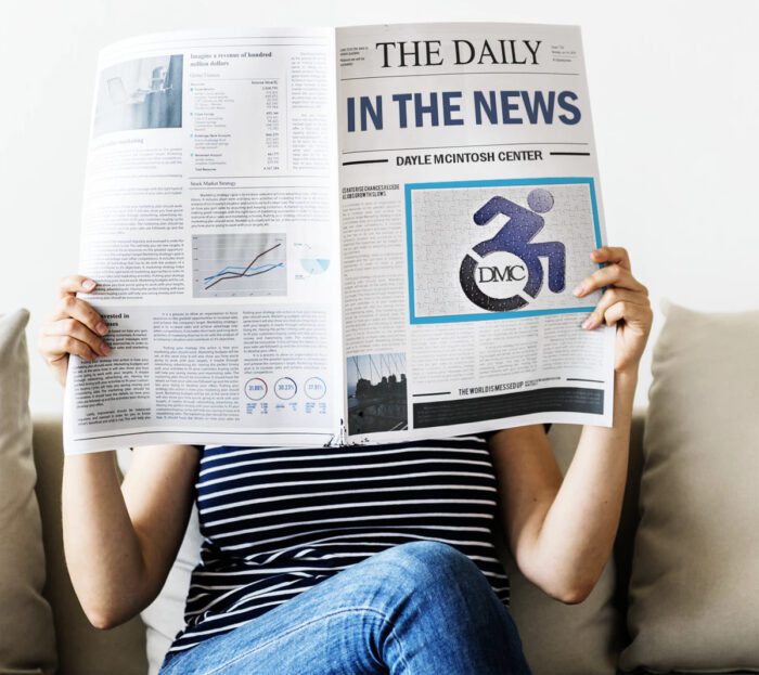 a person sitting on a sofa in striped T-shirt and jeans reading a newspaper holding it up in front of their face. The front of the newspaper says THE DAILY IN THE NEWS. Sub-headline is Dayle McIntosh Center with big photo of logo.