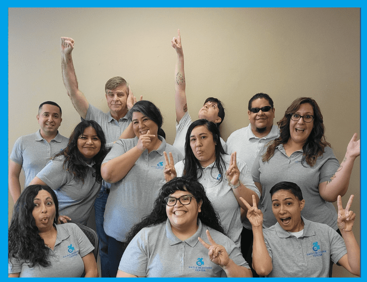 Pictured, The Dayle McIntosh Team representing a beautiful group of diverse people are all wearing staff DMC shirts in grey with blue wheelchair logo. Some team members are making peace signs, others pointing, all are joyful and jubilant with their expressions.