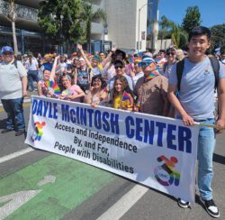 Pictured is the Dayle McIntosh Center team and volunteers smiling and celebrating during the 2023 Orange County Pride Parade. They are representing the organization and holding the banner which has the DMC logo in progressive pride colors with the text in blue and black across the banner reading “Dayle McIntosh Center – Access and Independence By, and For, People with Disabilities.