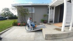 Person in wheelchair wearing baseball cap/hoodie using home modification ramp for home access from front porch