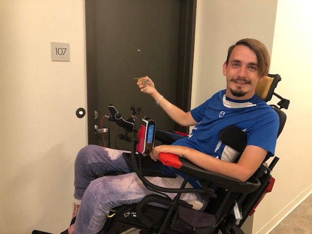 Pictured, consumer with disabilities, wearing a blue shirt and jeans with home keys in-hand, seated in a wheelchair experiencing a successful transition, returning back into the community to their home