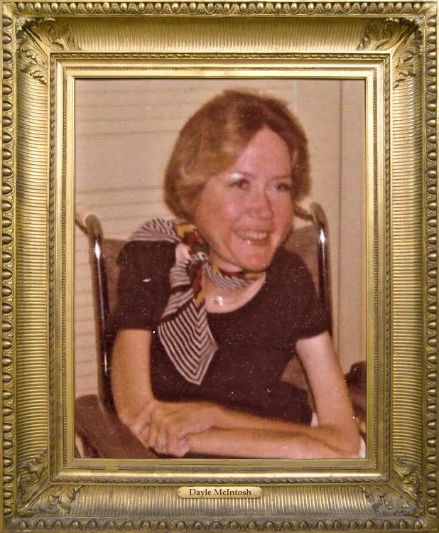 Pictured, DMC’s Founder, Dayle McIntosh smiling. She has short blonde hair and has a brown shirt and printed scarf. The photo is in a gold antique frame.