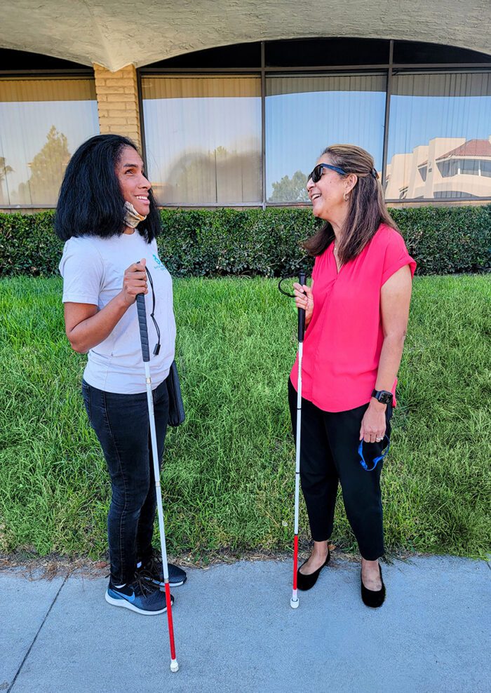 Danyelle and Socorro standing outside and facing each other as they talk. Both have pleasant smiles and are holding a white cane