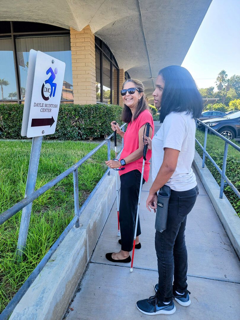  DMC Team Members Socorro and Danyelle using their white canes and walking up the side ramp of DMC's building. They are both facing a sign which has DMC's logo and a bold arrow pointing to the right