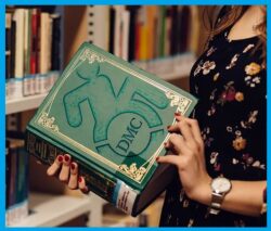 Person in a library hall with books all around, holding an encyclopedia-thick green book with The Dayle McIntosh logo on the cover featuring a gold antique frame to indicate it is an old book.