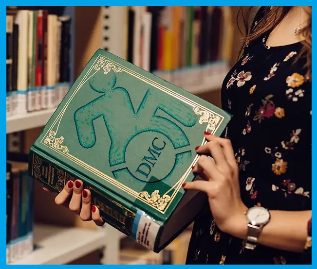  Person in a library hall with books all around, holding an encyclopedia-thick green book with The Dayle McIntosh logo on the cover featuring a gold antique frame to indicate it is an old book.