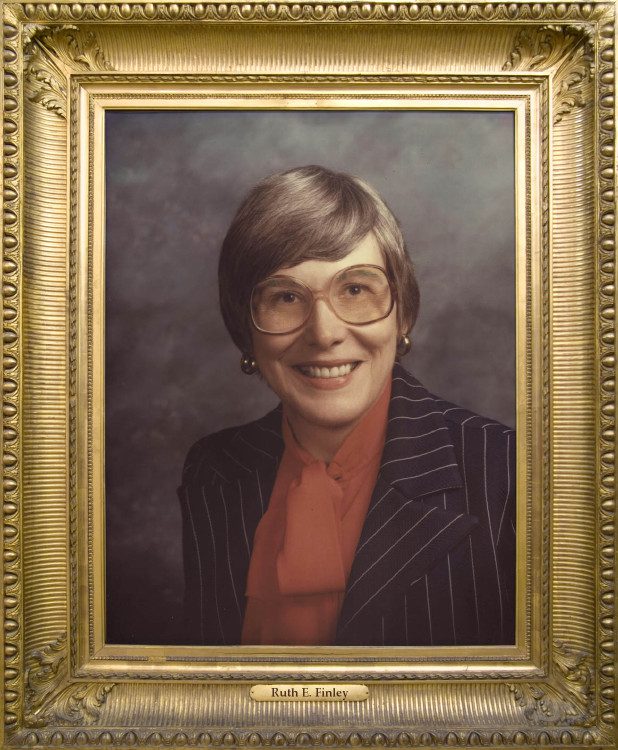 Photo of Ruth Finely wearing a black pinstriped suit with a red chiffon blouse. Ruth has ash-blonde hair, and is wearing large amber-tinted round rimmed eyeglasses, gold earrings