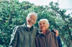 Older Adults - couple smiling outside in front of green trees and bushes.