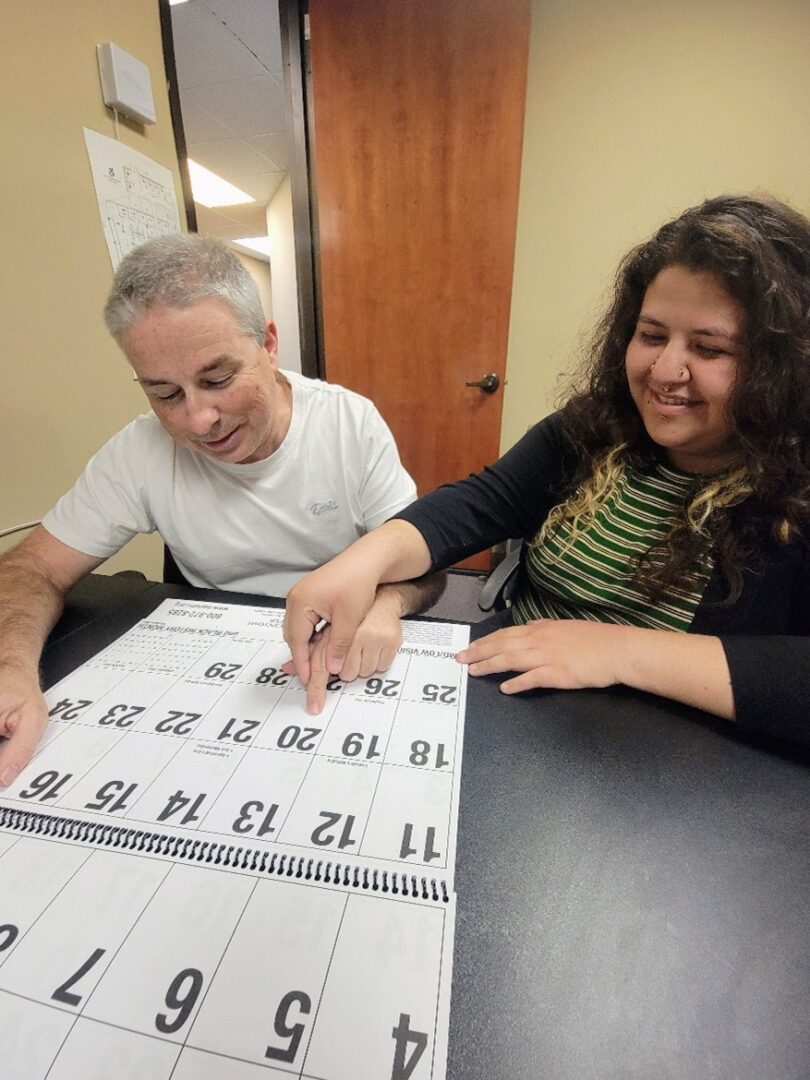 DMC Team Members Paul and Wendy demonstrating the use of a large print calendar. Wendy is guiding Paul’s hand to locate a date.