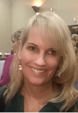 Pictured Suzanne Allee DMC's Board Member At-Large has blonde shoulder length-hair, dangling snowman earrings and is wearing a collared forest green blouse. She is smiling.