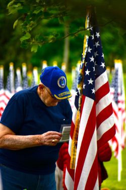 Veteran in blue shirt and blue Veterans cap standing in front of an American flag.