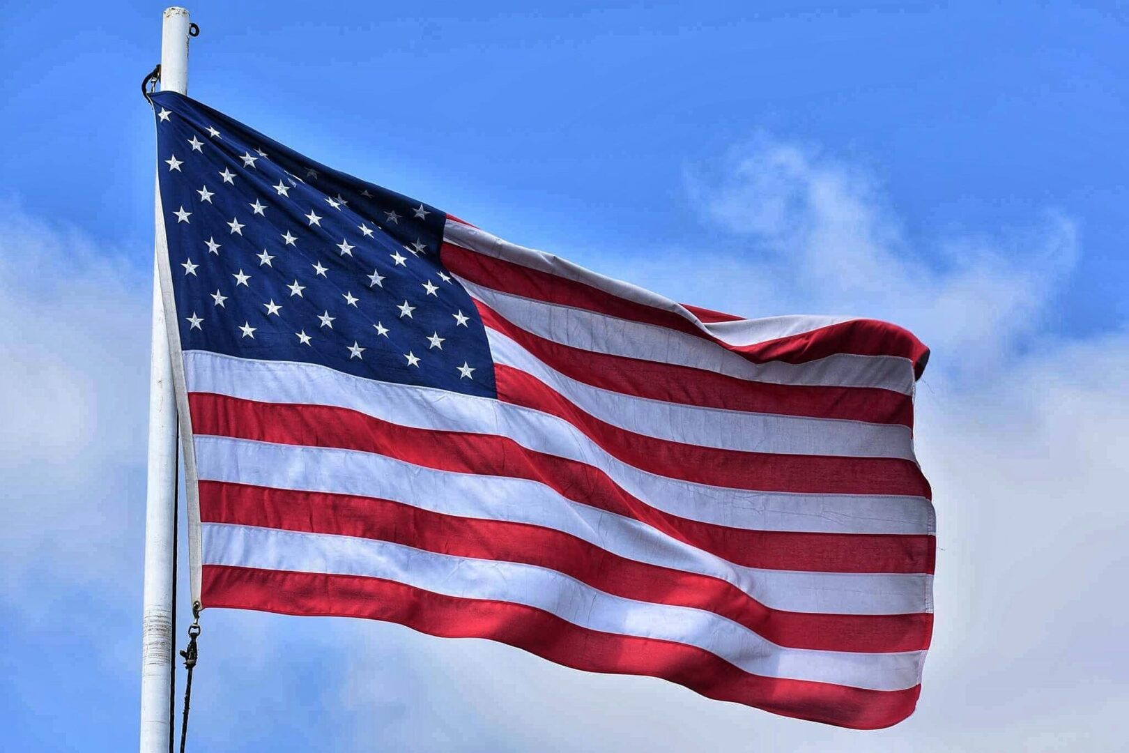 U.S. Flag red and white stripes and white stars on dark blue square in upper left corner on flag pole waving in the blue sky with white clouds.