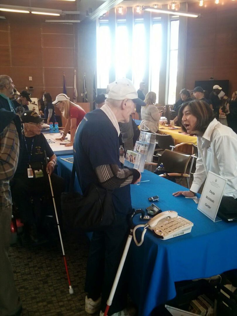 Pictured, Left: DMC’s retired Lead Older Individuals Who Are Blind (OIB) Advocate, Socorro Arroyo-Merchain is behind a table with a DMC resources speaking with a Veteran asking about one of the AT devices