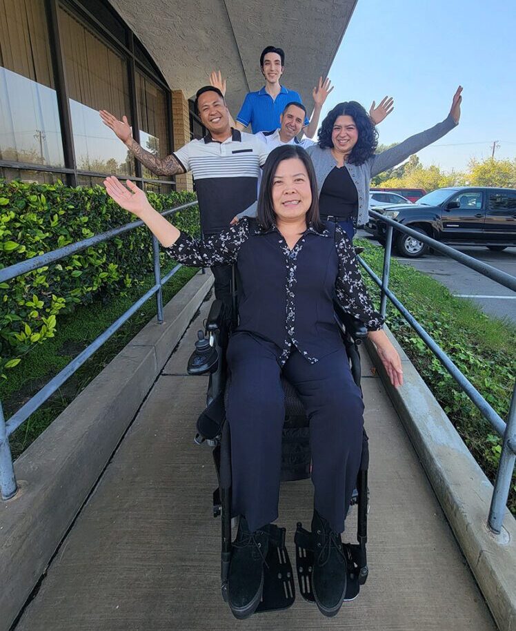 DMC Admin Team outside on the accessible ramp in front of the DMC. They are smiling with arms up and out, ready to help wherever it is needed! Front to back - Christine Nguyen, Jackie Castro, Michael Susi, Miguel Escarzaga, and Peter Humphreys.