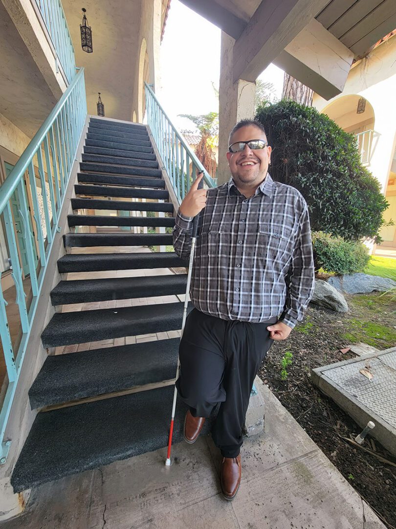 Pictured, DMC Youth Community Liaison and State Independent Living Council, Alan Cruz standing in front of outside open stairwell on a sunny day at DMC, smiling in blue plaid shirt.