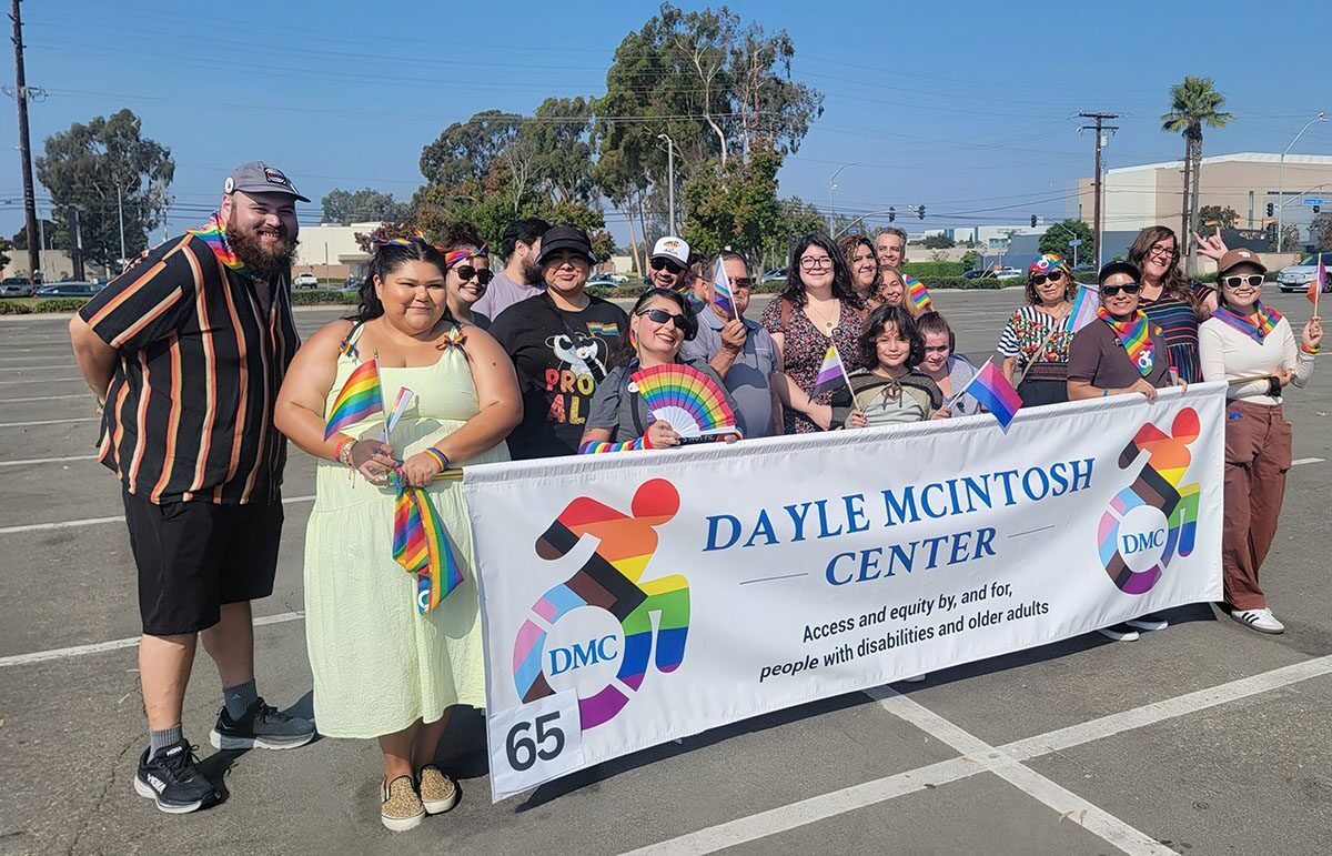 A group photo featuring our team, consumers, friends, and family proudly standing by our DMC banner, showcasing our logo in the colors of the progressive pride flag alongside our mission statement.