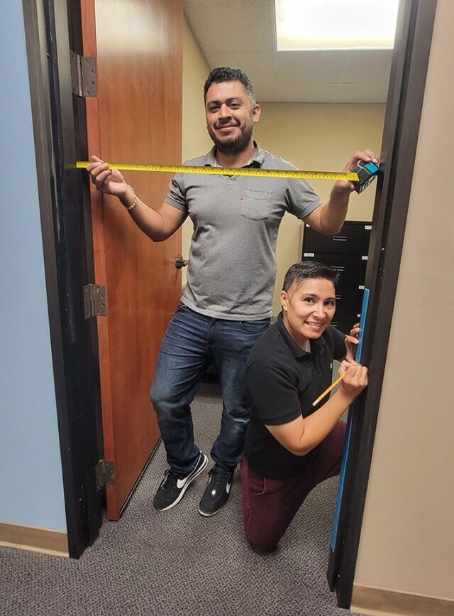  Pictured Home Access Team Ivan with measuring tape and Ana marking measurements with pencil on fame, as both are ensuring that measuring door dimensions are noted accurately to create access. 