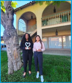Deaf Services Team, Pamela, left holding a heart light and Doryna, right, holding an ASL ILY light, standing outside outside smiling on green grass underneath a beautiful tree..