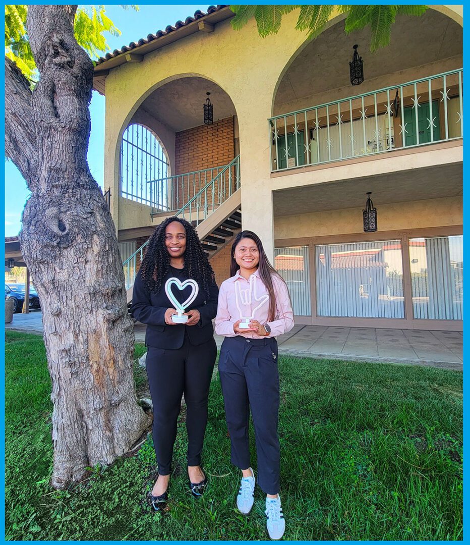 Deaf Services Team, Pamela, left  holding a heart light and Doryna, right, holding an ASL ILY light, standing outside outside smiling on green grass underneath a beautiful tree..