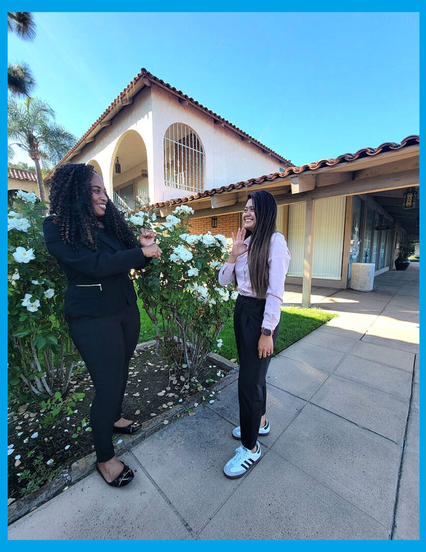 Pictured Deaf Services team communicating outside. Left, Pamela signing to Doryna, with Doryna reacting. They are standing in front of white roses in the garden.