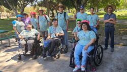 Pictured, 2024 DMC Youth Summer Academy – A celebration of Peace, Love and Flower wreaths. Everyone is outside smiling in a park with lots of trees in the background; Lead Youth and Information Coordinator, Nelly is smiling holding up two fingers as a peace sign, leading the summer celebration!