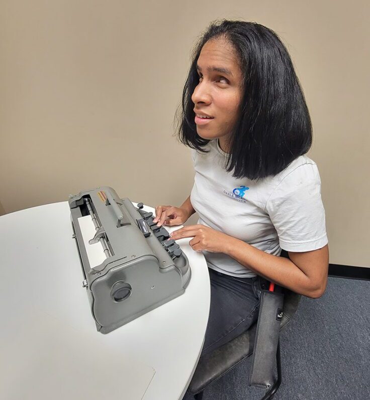 Pictured Team DMC Member, Danyelle sitting at table, using the Perkins Brailler machine to write notes in Braille.