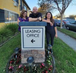 Pictured: Housing Team’s Marisol, Adam and Lizeth standing outside over an apartment Leasing Office Sign which is propped on a brown brick post, surrounded by outdoor ground lights and pink and red flowers with grass. A yellow apartment building and trees are behind them as they smile about providing housing options to consumers.