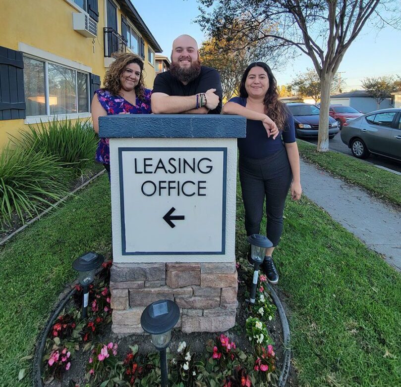 Pictured: Housing Team’s Marisol, Adam and Lizeth standing outside over an apartment Leasing Office Sign  which is propped on a brown brick post, surrounded by outdoor ground lights and pink and red flowers with grass. A yellow apartment building and trees are behind them as they smile about providing housing options to consumers. 