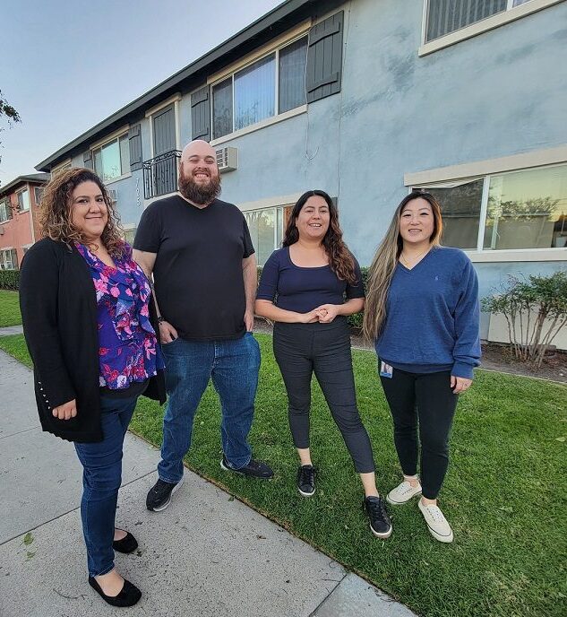 Pictured DMC Housing Team L-R: Marisol, Adam, Lizeth and Ruth standing outside in front of apartment building, smiling. 