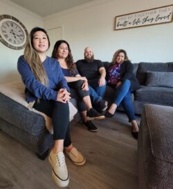 Pictured Team DMC Housing Services Team L-R: Ruth, Lizeth, Adam and Marisol sitting on sofa in rental property. A clock is featured on the wall on the left and a sign on the all on the right that reads: “And so together they built a life they loved.”