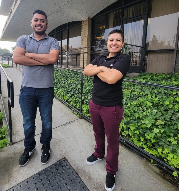 Pictured Ivan and Ana, DMC’s Home Access Team standing outside DMC on access ramp outside of DMC building, with smiles and arms crossed.